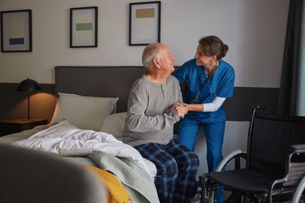 A caregiver provides in-home Parkinson's care to her client, offering mobility support in getting out of bed.
