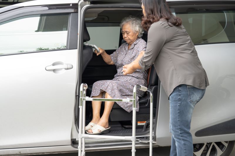 A caregiver helps an elderly woman get into a vehicle.