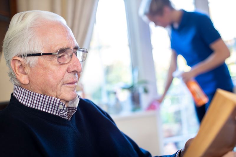 An elderly man reads a book while his caregiver cleans his home.